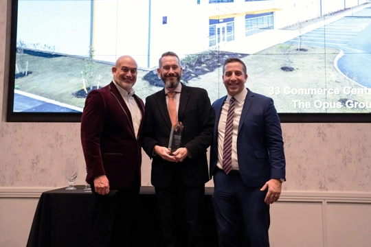 three men wearing suits with the man in the center holding an award